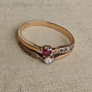 May include: A gold ring featuring a split shank design with a central round red ruby and a round clear diamond set vertically. The band is accented with small embedded stones along the sides, creating a detailed and structured appearance.