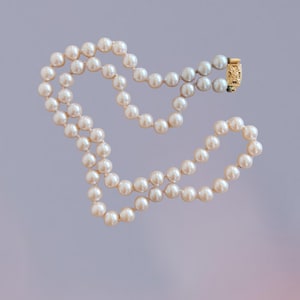 May include: A single strand of cream-colored pearls arranged in a loose, circular shape. The necklace features a rectangular, gold-toned metal clasp with an intricate, embossed floral pattern that secures the two ends of the pearl strand together.