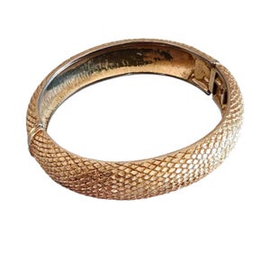 May include: A gold-toned hinged bangle bracelet featuring a textured, cross-hatched surface pattern that resembles scales. The bracelet has a smooth, polished interior and a secure metal clasp mechanism visible on the side of the circular band.