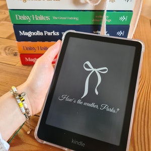 May include: A black and white Kindle e-reader with a white bow and the text "How's the weather, Parks?" on the screen. The e-reader is on a wooden surface with a stack of books behind it.