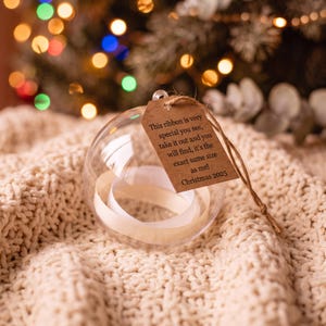 Puede incluir: Un adorno esférico transparente que contiene una cinta blanca, unido a una etiqueta de papel marrón con cordel. La etiqueta presenta el texto: This ribbon is very special you see, take it out and you will find, it's the exact same size as me! Christmas 2025.
