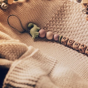 May include: A baby's dummy clip with wooden beads and a green dinosaur charm. The clip features the name "ROHAN" spelled out in wooden letter beads, and the name "CODY" is also visible. The dummy clip is on a cream-coloured knitted blanket.