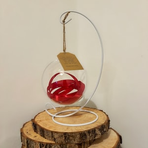 May include: A clear glass bauble filled with red ribbon, hanging from a white metal stand. A small tag with text is attached to the bauble. The stand sits on a stack of wooden slices, creating a rustic display.