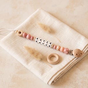 May include: A dummy clip featuring a string of pink and beige silicone beads, a flower-shaped bead, and white cube beads spelling the name KATIE. The clip includes a wooden fastener and a small wooden ring, all arranged on a folded cream cloth.