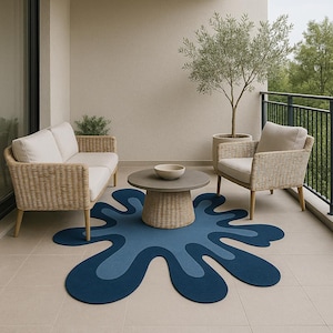 Op de afbeelding: Een balkonsc&egrave;ne met een blauw, abstract gevormd tapijt. Rotan meubels met cr&egrave;mekleurige kussens, een ronde salontafel en een olijfboom in pot staan op de betegelde vloer. Het tapijt heeft twee blauwtinten.