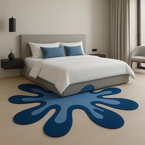 Op de afbeelding: Een moderne slaapkamer met een grijs bed, wit beddengoed en blauwe kussens. Een groot, abstract blauw tapijt in een spattendesign ligt op de lichte houten vloer. De kamer heeft neutrale muren en een minimalistische esthetiek.