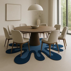 Op de afbeelding: Een moderne eethoek met een ronde houten tafel met een donkerbruine voet, omringd door acht beige gestoffeerde stoelen met houten poten. Een blauw, abstract gevormd tapijt ligt onder de tafel. Een witte hanglamp hangt erboven.