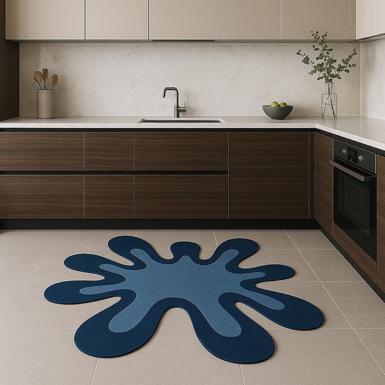 Op de afbeelding: Een blauw, spatvormig tapijt met een lichtblauw midden ligt op een betegelde vloer in een moderne keuken. Donkere houten kasten en lichte werkbladen vormen een neutrale achtergrond. Een roestvrijstalen kraan en oven zijn zichtbaar.