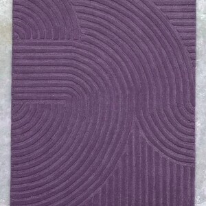 May include: A purple rug with a geometric pattern of curved lines. The rug has a textured surface and a modern design.