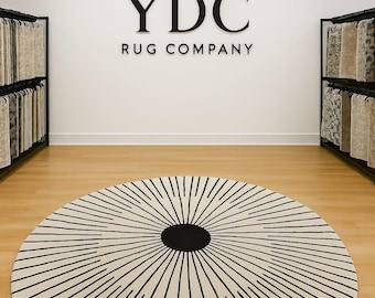 May include: A round area rug featuring a cream-colored base with a black circular center and radiating black lines extending to the edge. The rug is displayed in a showroom with black metal racks holding various rug samples and the text YDC RUG COMPANY.