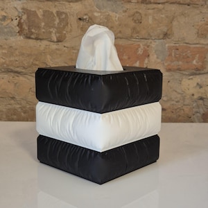 Könnte beinhalten: Ein quadratischer Taschentuchspender mit modernem Design. Die Box besteht aus drei übereinander gestapelten Schichten: schwarz, weiß und schwarz. Weiße Taschentücher sind oben sichtbar. Die Box steht auf einer weißen Oberfläche vor einer Backsteinmauer.