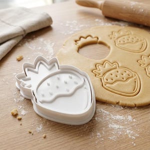 May include: A white plastic strawberry-shaped cookie cutter beside a sheet of dough with strawberry imprints. A rolling pin and a folded beige cloth are in the background. The scene is set on a light brown wooden surface.