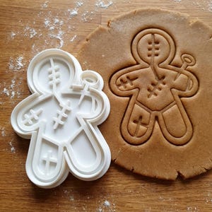 May include: A white plastic gingerbread man cookie cutter next to a cut-out gingerbread man shape in dough. The cutter has a detailed design with stitched accents and a small tool. The dough is light brown, and the background is a wooden surface.
