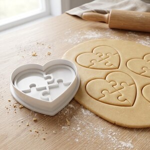 Puede incluir: Un cortador de galletas blanco en forma de corazón con un diseño de rompecabezas junto a galletas cortadas sobre una superficie de madera. Un rodillo y un paño están en el fondo. Las galletas están cortadas en forma de corazón con diseños de piezas de rompecabezas.