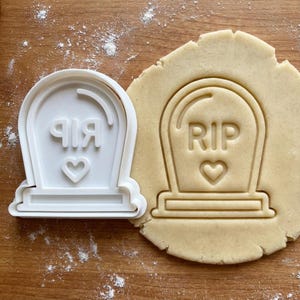 May include: A white plastic tombstone-shaped cookie cutter with the letters "RIP" and a heart. Next to it is a tombstone-shaped imprint in a piece of dough. The cutter is for making cookies.