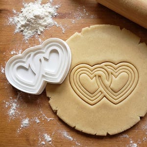 Puede incluir: Juego de cortadores de galletas de plástico blanco en forma de dos corazones entrelazados, junto a un trozo de masa con las formas de corazón presionadas. Harina espolvoreada sobre la superficie de madera, con un rodillo en el fondo. Tema de repostería y San Valentín.