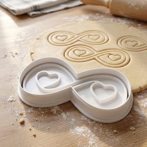 Könnte beinhalten: Weißer Ausstecher in Form eines Unendlichkeitszeichens mit einem Herz im Inneren. Der Ausstecher befindet sich neben einem ausgerollten Teig mit eingepressten Unendlichkeits- und Herzformen. Ein Nudelholz und ein gefaltetes Tuch sind im Hintergrund.