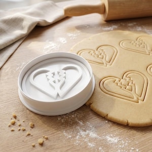 May include: A white heart-shaped cookie cutter with a unique design sits next to rolled-out dough with heart imprints. A wooden rolling pin and a cloth are in the background. The scene suggests baking and Valentine's Day.