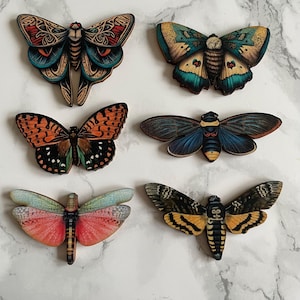 May include: Six wooden butterfly and moth ornaments featuring intricate details and vibrant colours. The butterflies are painted with realistic patterns and colours, including orange, blue, green, and red.
