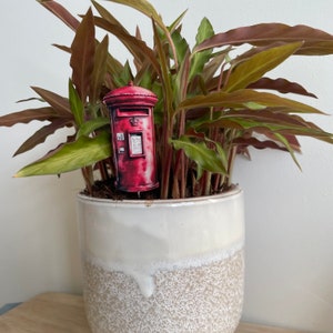 Può includere: Una cassetta delle lettere rossa e dorata con una corona in cima è posizionata in un vaso di ceramica bianco con una glassa marrone macchiata. Il vaso è pieno di una pianta verde a foglie con steli rossi.