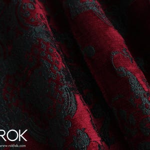 May include: Close-up of a deep red fabric with a black, ornate damask pattern. The fabric is folded, showing the texture and depth of the design. The word "ROK" is visible in white text at the bottom left.
