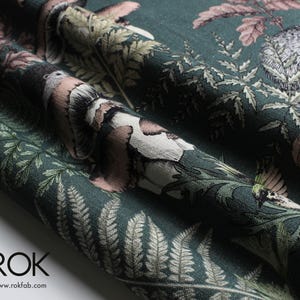 May include: Dark teal fabric with a detailed forest print featuring birds, foliage, and woodland creatures. The design incorporates shades of brown, pink, and green. The fabric is folded, showing the texture and pattern. The logo "ROK" and website address are visible.