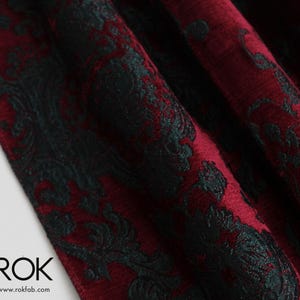 May include: Close-up of a burgundy fabric with a dark green, ornate damask pattern. The fabric is folded, showing texture and depth. The logo "ROK" and website address are visible in the lower left corner.