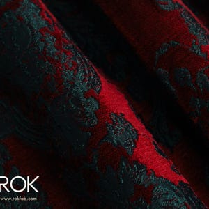 May include: Close-up of a textured fabric with a damask pattern. The fabric features a deep red background with a contrasting dark teal floral design. The word "ROK" and website address are visible in the lower left corner.