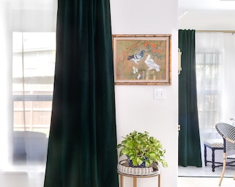 May include: A room with white walls and dark green velvet curtains. A framed painting of two birds is on the wall. A small gold side table with a potted plant sits in the corner of the room.
