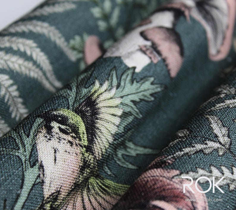 May include: Close-up of a fabric with a detailed botanical print. The design features birds, flowers, and foliage in shades of green, pink, and cream against a dark teal background. The fabric is folded, showing the texture and pattern.