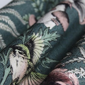 May include: Close-up of a fabric with a detailed botanical print. The design features birds, flowers, and foliage in shades of green, pink, and cream against a dark teal background. The fabric is folded, showing the texture and pattern.