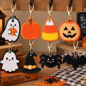 May include: A set of eight embroidered Halloween keychains featuring a white ghost holding a pumpkin, an orange pumpkin, a candy corn, a jack-o'-lantern, a second ghost, a black witch hat, a black spider, and a black bat, all with gold clasps.