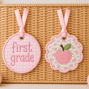 May include: Two embroidered fabric tags with pink ribbon loops. One circular tag features a light pink gingham pattern with the text first grade in pink thread. The other scalloped tag displays a floral print with a pink apple and green leaf.