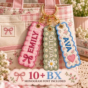 May include: Three rectangular scalloped fabric keychains with gold-tone metal clasps attached to a pink and white floral bag. The tags feature embroidered names EMILY, OLIVIA, and AVA with decorative bow, daisy, and heart motifs in pink, green, and blue.