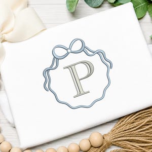 May include: A white shirt featuring an embroidered design of a light blue ribbon tied into a bow, which forms a circular frame around the capital letter P in a light gray color. The embroidery is centered on the front of the garment.