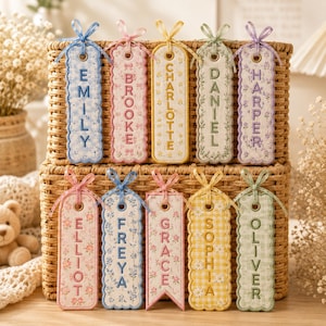 May include: Ten rectangular fabric bookmarks with scalloped edges and ribbon ties at the top. Each bookmark features a unique floral or gingham pattern in pastel colors and is embroidered with a name: EMILY, BROOKE, CHARLOTTE, DANIEL, HARPER, ELLIOT, FREYA, GRACE, SOPHIA, and OLIVER.