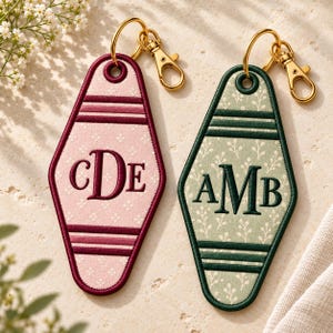 May include: Two diamond-shaped motel-style keychains with gold-tone lobster clasps. The left keychain is pink with a small floral pattern and dark red embroidery reading CDE. The right keychain is sage green with a leaf pattern and dark green embroidery reading AMB.