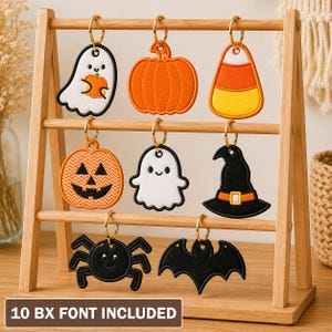 May include: A wooden display stand holding eight embroidered Halloween-themed keychains, including a ghost, pumpkin, candy corn, jack-o'-lantern, witch hat, spider, and bat. A brown rectangular sign at the bottom reads 10 BX FONT INCLUDED.
