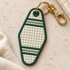 May include: A diamond-shaped motel-style keychain featuring a light sage green and white gingham pattern. The keychain is bordered with dark green stitching and includes two horizontal dark green stripes. It is attached to a gold-tone metal lobster clasp.