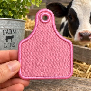May include: A bright pink embroidered patch shaped like a livestock ear tag with a circular hole at the top. The patch features a dense, uniform thread texture throughout its surface and a thick, raised border stitched in the same vibrant pink color.