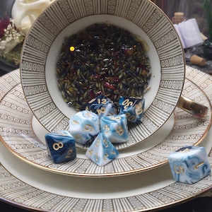 Könnte beinhalten: Ein Porzellan-Tassenset mit Untertasse, verziert mit einem braun-weißen Streifenmuster und Goldrand. Die Tasse ist mit getrocknetem Lavendel und Blütenblättern gefüllt und enthält mehrere blau-weiß marmorierte Polyederwürfel mit goldenen Zahlen.