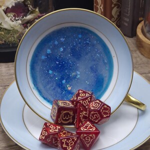 Könnte beinhalten: Ein hellblaues Teetassen- und Untertassenset mit Goldrand, gefüllt mit einer durchscheinenden blauen Harzmischung mit Glitzer. Eine Ansammlung dunkelroter polyedrischer Würfel mit goldfarbenen filigranen Zahlen liegt in der Tasse und auf der Untertasse.