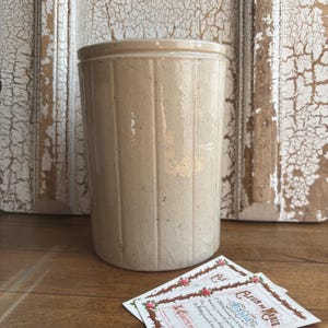 May include: A cylindrical beige stoneware crock featuring vertical ribbed detailing along its exterior surface. The vessel has a smooth, glazed finish with subtle speckling throughout the clay body and a slightly raised rim at the top edge.