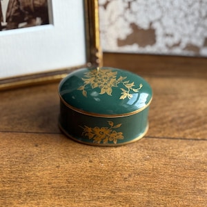 May include: A small, round, green trinket box with a gold floral design. The box has a gold rim and lid. The box is on a wooden surface. A framed photograph is in the background.