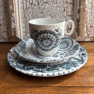 May include: A white teacup with a handle sits on a matching saucer and a larger plate. The teacup and plates feature a blue floral pattern. The set is displayed on a wooden surface, suggesting a vintage aesthetic.