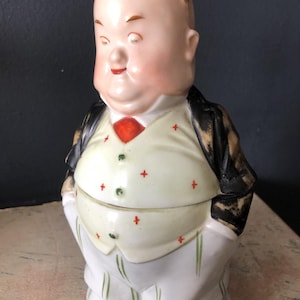 May include: A vintage ceramic figurine of a stout man in a suit. The figure has a bald head with small horns, a red tie, and a white vest with red cross accents. The suit jacket is black with gold accents.