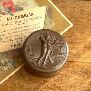 May include: A round, dark brown metal powder compact featuring a relief sculpture of two figures dancing on the lid, surrounded by a textured, radiating sunburst pattern. The lid surface is detailed with fine, concentric, etched lines.