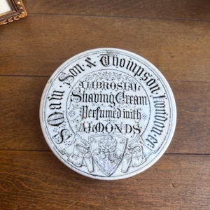 May include: A circular, white ceramic shaving cream container with black text. The text reads "Wm. Son & Thompson, London" around the edge, and "Ambrosial Shaving Cream, Perfumed with Almonds" in the centre. A floral crest is in the middle.