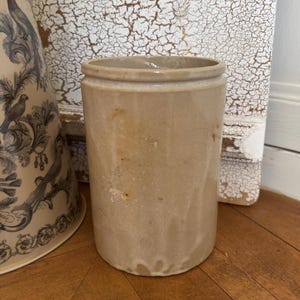 May include: A cylindrical stoneware crock with a smooth, light tan glaze and a subtle, mottled texture. The vessel features a simple, straight-sided design with a slightly raised rim at the top, showcasing a neutral, earthy tone and a sturdy, functional form.