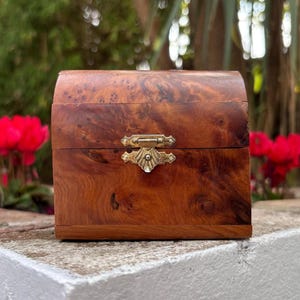 May include: A small, arched-top wooden box featuring a rich, swirling burl wood grain pattern in shades of medium to dark brown. The box is secured with a decorative, gold-toned metal latch centred on the front, showcasing a detailed shell motif.